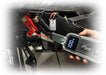 A hand holds a gray and black iMoto Super digital battery monitor connected to a motorcycle battery with red and black clamps, shown against a blurred outdoor background.