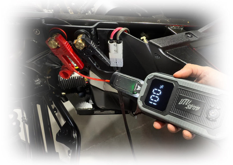A hand holds a gray and black iMoto Super digital battery monitor connected to a motorcycle battery with red and black clamps, shown against a blurred outdoor background.